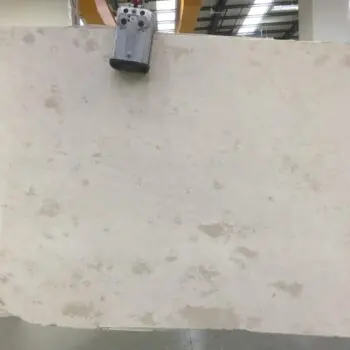 The MA009 - Travertine Classico is a large rectangular light beige slab with subtle natural patterns, suspended in an industrial warehouse by a mechanical clamp.