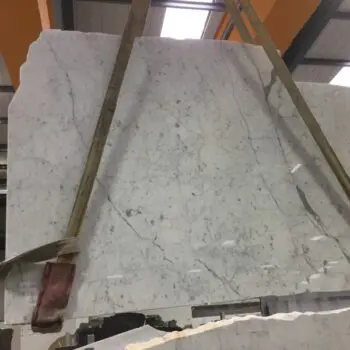 A large slab of MA185 - Carrara Bianco Gioia, featuring white marble with grey veining, is suspended by two straps inside an industrial warehouse.