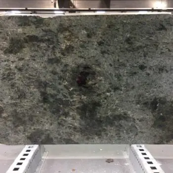 A large rectangular slab of Labradorite Semi-Precious Stone, featuring dark speckles, is displayed upright on a metal stand in an indoor industrial setting.