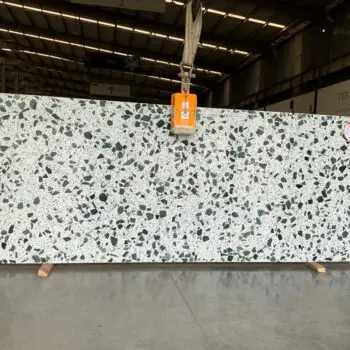 A large TE112 terrazzo slab featuring black, white, and grey stone chippings is suspended by a crane inside an industrial warehouse.